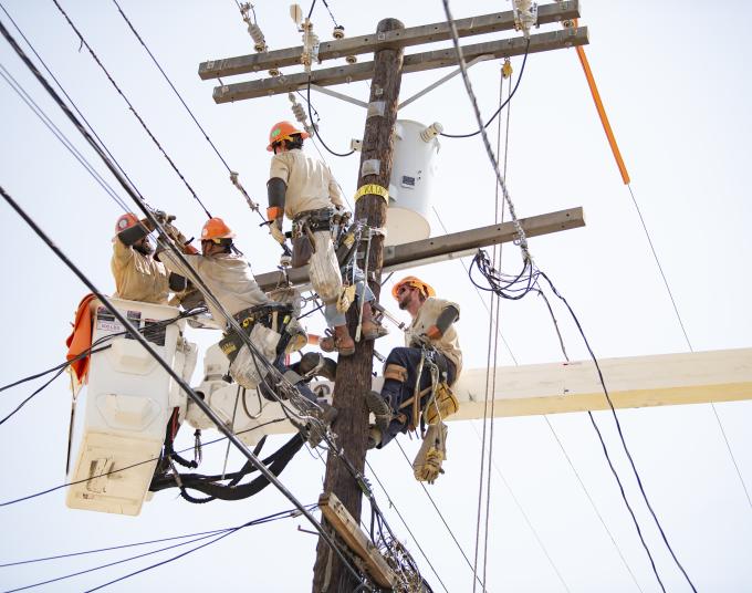 Four electrical distribution mechanics replace a transformer high up on a power pole. One of them is working from a hydraulic lift bucket and the three remaining are harnessed to the pole.