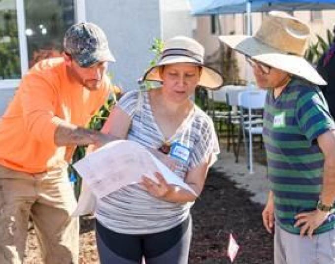 Three people in the front yard of a house. One man points at a sheet of paper while a second man and a woman look at what he’s pointing.