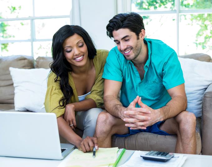 Smiling couple sitting on a couch in front of a table that has a laptop, notepad, and calculator. The woman is holding a pen and pointing to the paper and the man is looking at the paper.