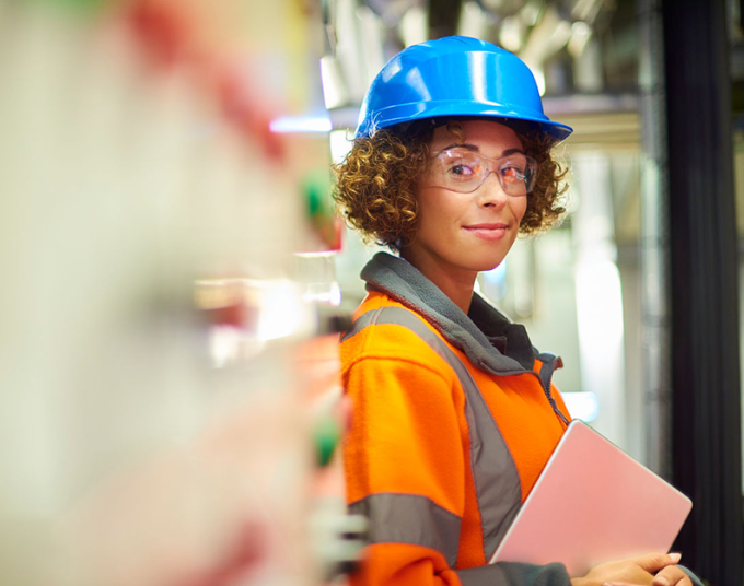 A woman in an orange jacket, blue hard hat, and safety glasses holds documents and smiles at the camera.