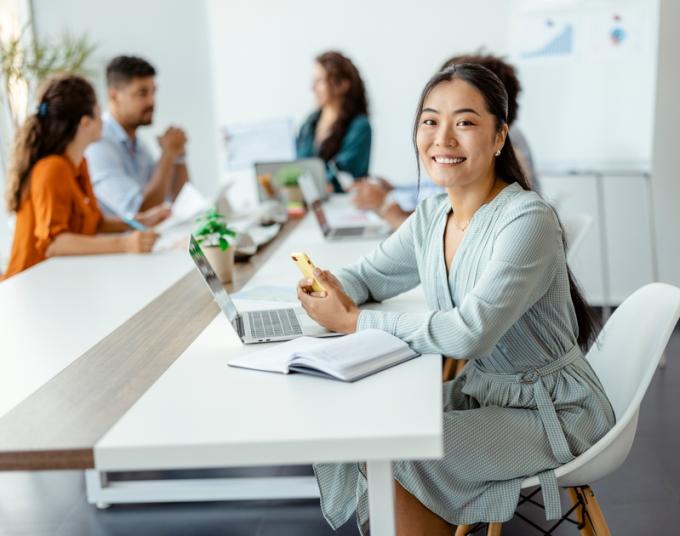 A smiling businesswoman sits in front of her laptop while working in a well-lit shared office space.