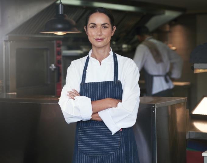 A smiling woman in an apron stands in a commercial kitchen.