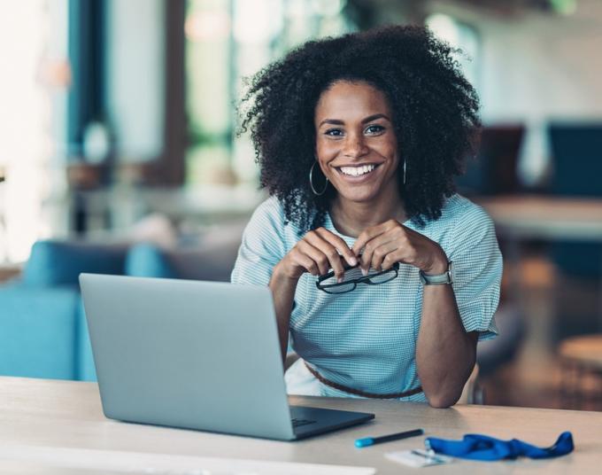 A woman smiles and holds her glasses while sitting in front of a laptop.