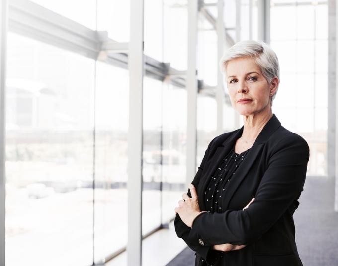 A businesswoman looks at the camera while standing in front of large windows.