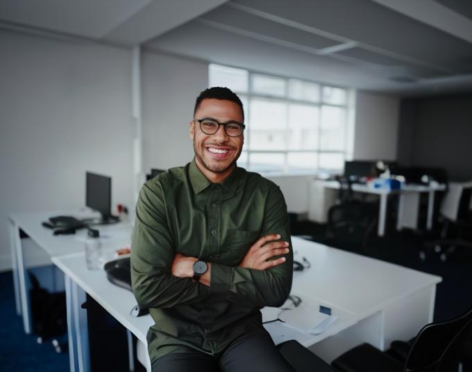 A smiling businessman sits on the edge of a workstation.