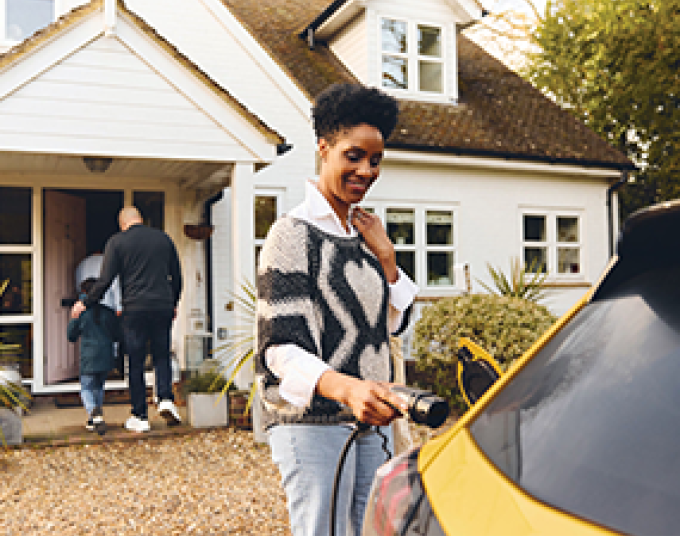 Woman is standing outside of a home attaching her charger plug to her yellow electric vehicle.