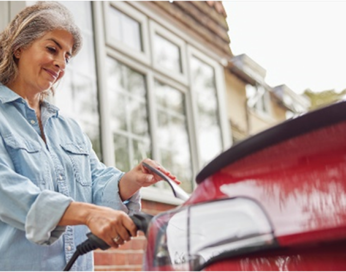 An older woman with grey hair is holding an electric vehicle charger cord to her red car.