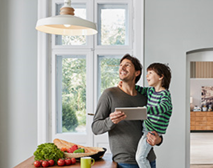 Smiling man carrying smiling kid while holding a tablet and looking up at the kitchen light. 