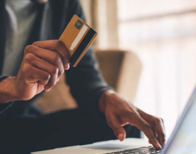 A person holding a gold credit card up while pressing a key on their laptop with their other hand. They are seated on a beige couch.