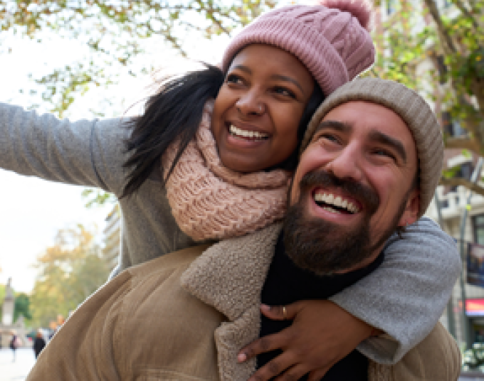 A smiling couple outside in nature wearing winter clothes. The woman is on top of the man’s back and is wearing a pink beanie and pink scarf. The man is holding the woman on his back and is wearing a tan sherpa coat and a tan beanie.