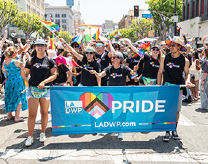 A group of people wave and march while holding a blue banner that reads, “PRIDE. ladwp.com” 