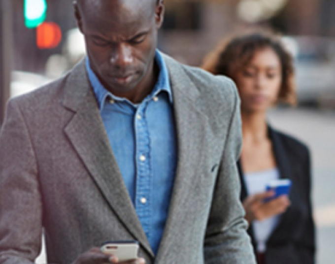 Man wearing a grey blazer with a blue jean button-up and a woman wearing a black blazer are standing outside. Both are looking at cell phones in their hands.