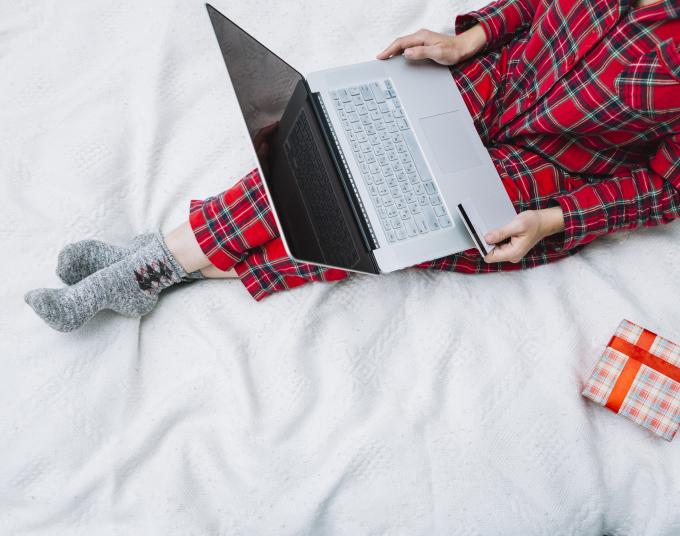 A person seated on a bed with a computer laptop resting on their legs.