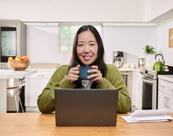 Smiling woman sitting at a kitchen table with a laptop and papers while holding a mug.