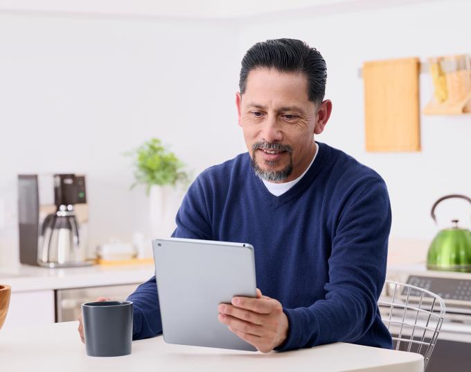 Man seated in kitchen looking at a computer tablet.