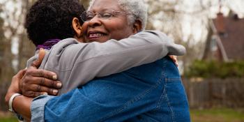 A grey-haired woman smiling as she’s hugging a child outside in a yard.