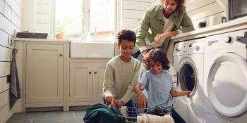 A man with curly hair is smiling and leaning against a white laundry machine, looking down at two kids. The two kids are smiling and taking things out of the laundry and putting it in a wire basket.