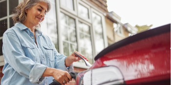 An older woman with grey hair is holding an electric vehicle charger cord to her red car.