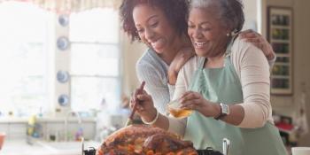 Two smiling women standing at a kitchen counter as one bastes a turkey.