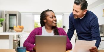 Woman is sitting at a kitchen table working on a laptop, looking up at a man standing next to her. The man is looking at her while holding papers.