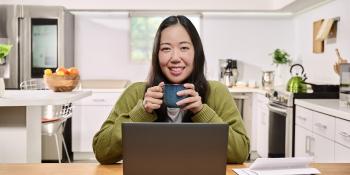Smiling woman sitting at a kitchen table with a laptop and papers while holding a mug.